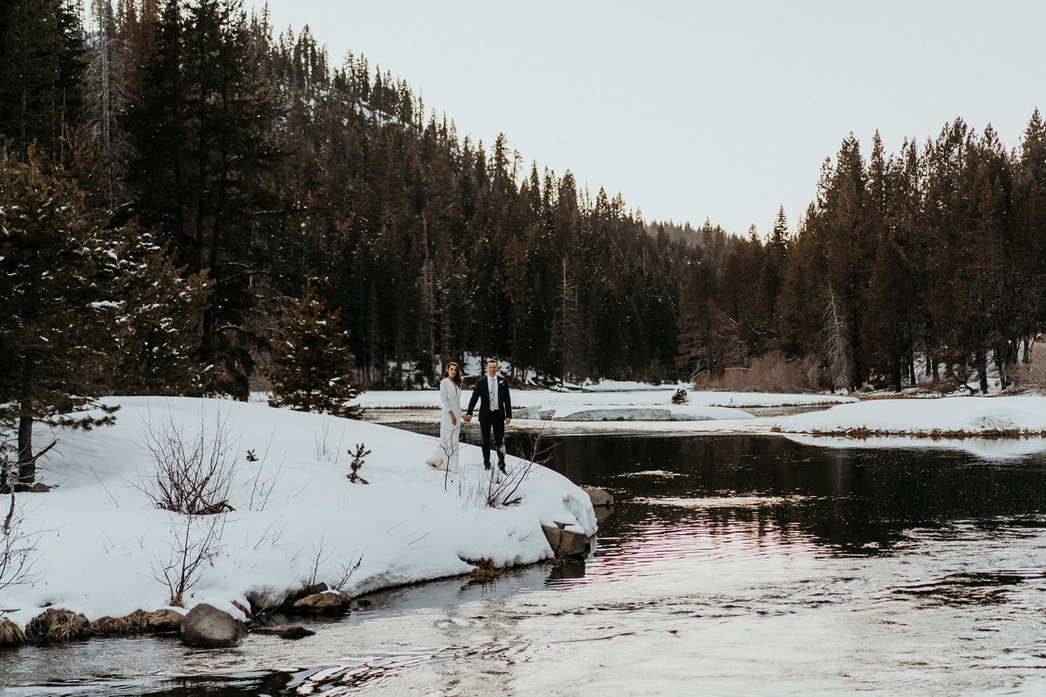 Bride and groom stand on a snowy bank next to a river during their elopement in Lake Tahoe, California.