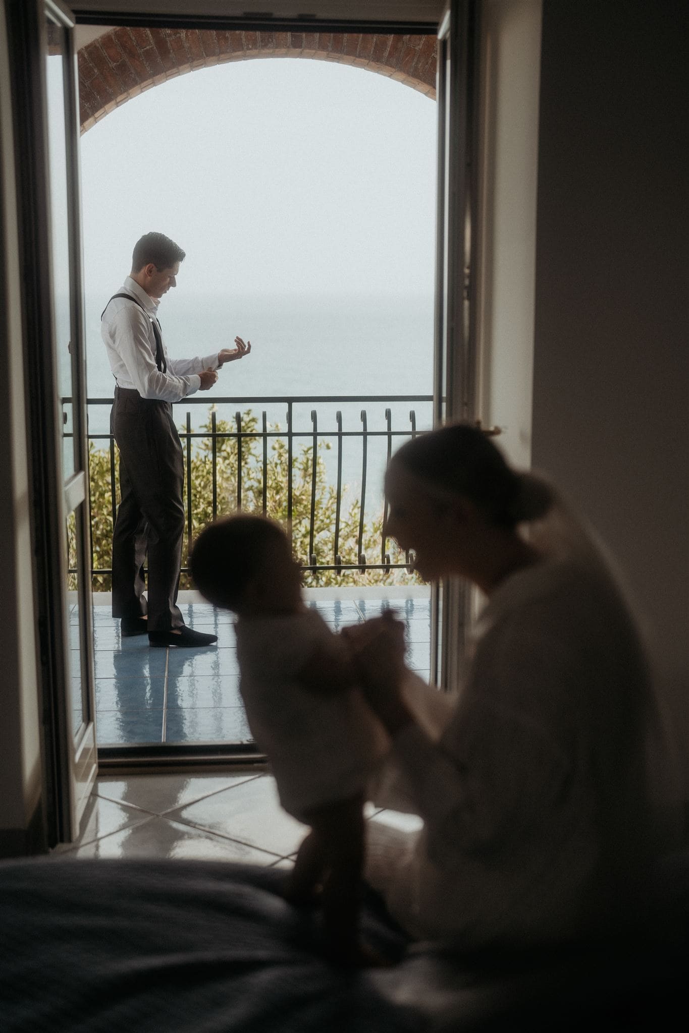 Bride plays with baby on the bed while groom gets ready on the patio