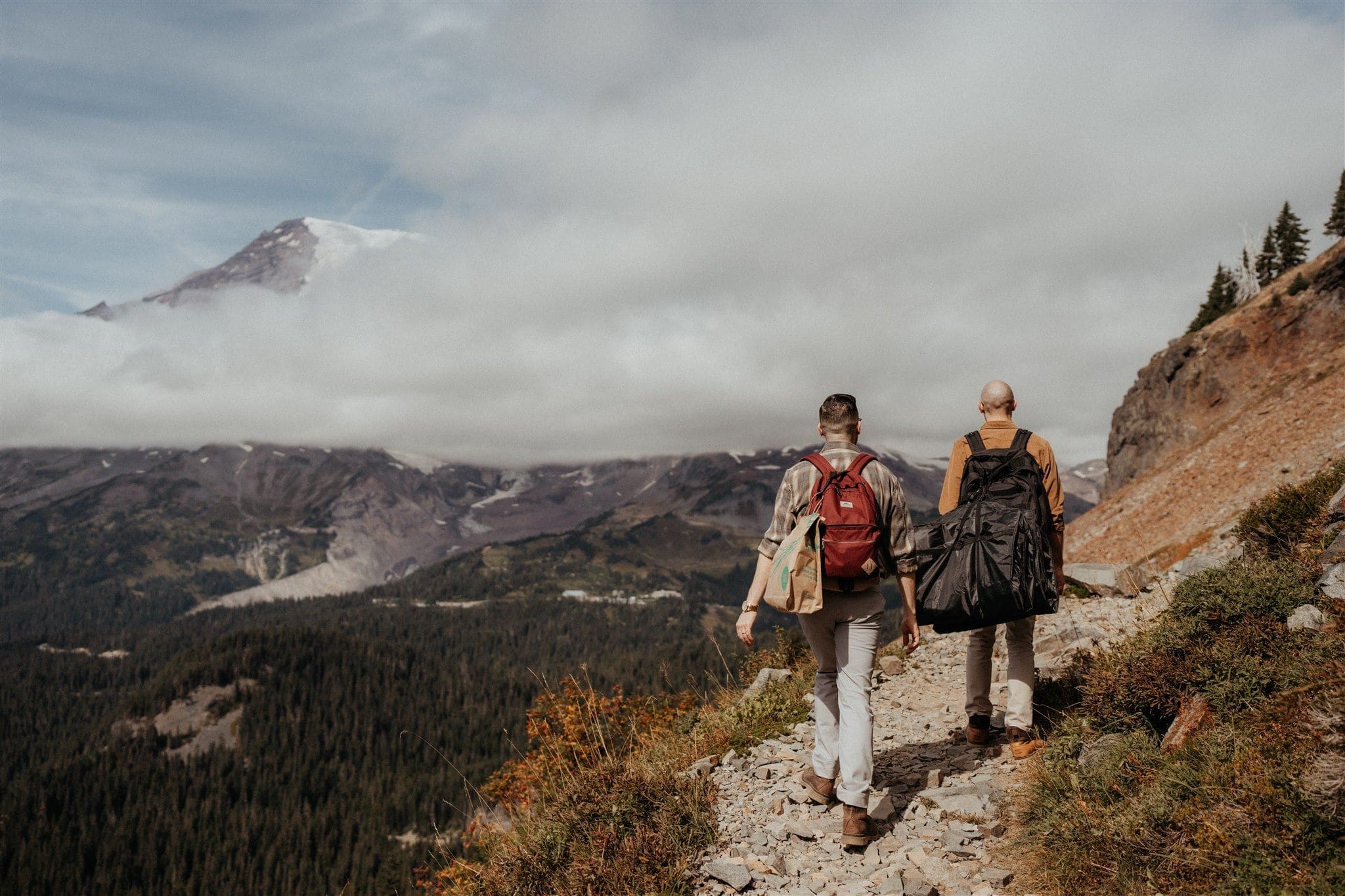 Two grooms hike with backpacks to their elopement in the mountains
