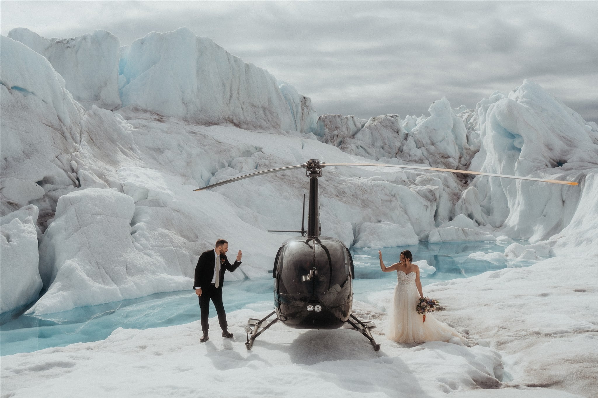 Bride and groom wave to each other through helicopter windows at their elopement in Alaska