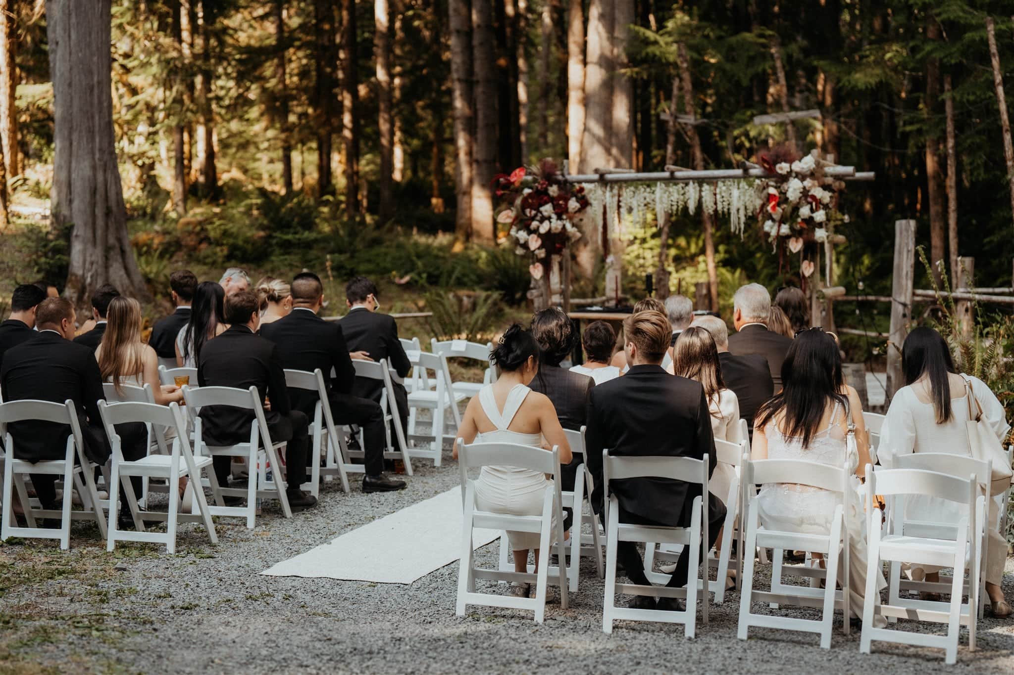 Guests sit in white wood chairs during an intimate wedding ceremony in the forest