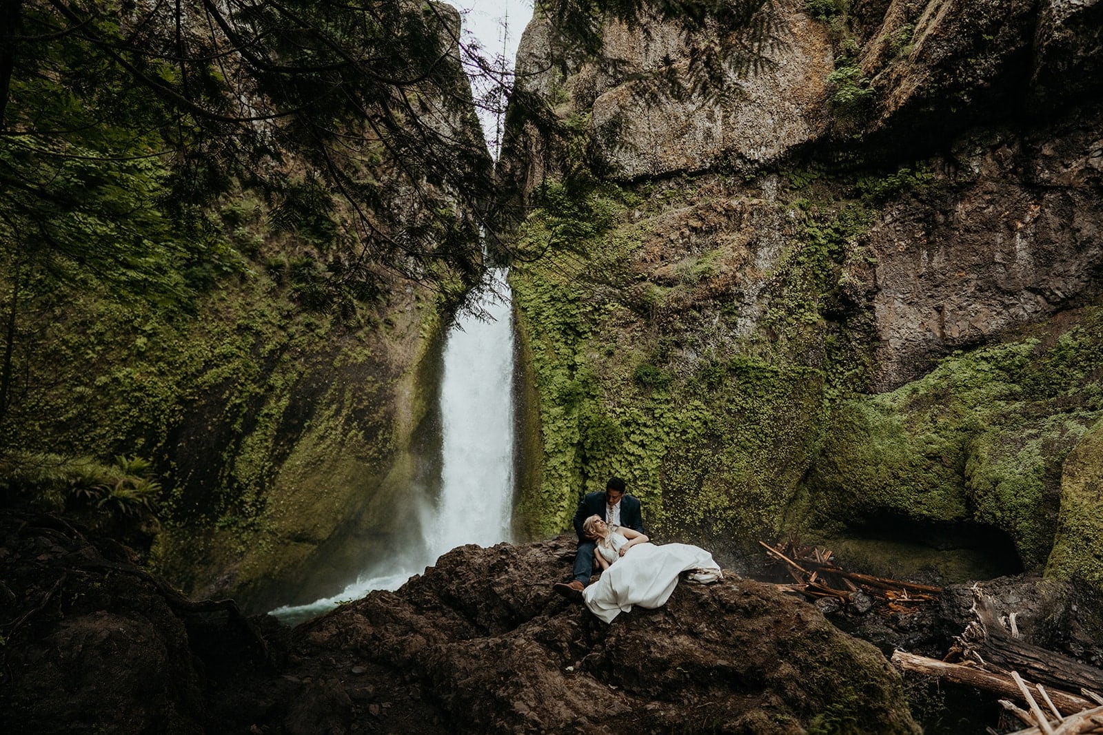 Bride and groom sit on a rock beneath a waterfall in the Columbia River Gorge.