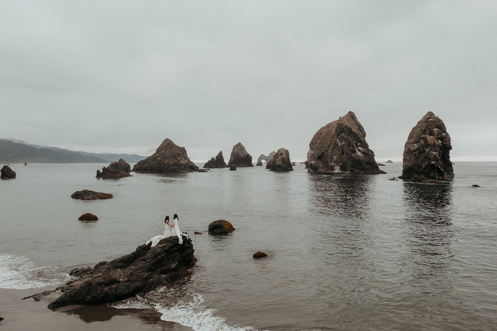 Two brides stand on a rock overlooking the ocean during their adventure elopement on the Oregon Coast.
