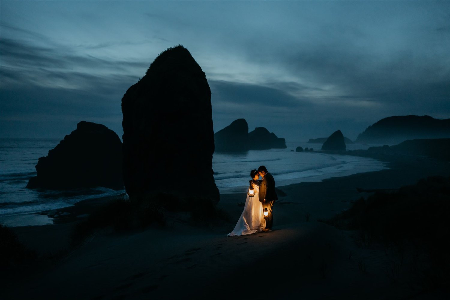 Two brides stand with their foreheads touching while holding lanterns during blue hour on the Oregon Coast.