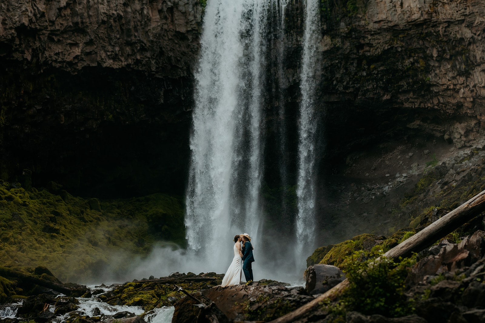 Two brides kiss while standing at the base of a waterfall during their Oregon elopement.