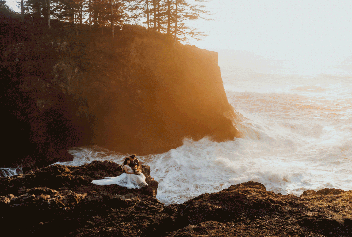 Two brides sit on the rocky cliffs of the Oregon Coast during their sunset adventure elopement.