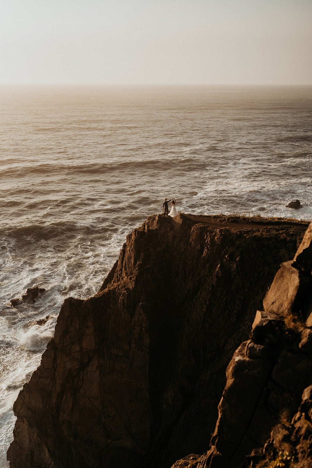 Two brides stand on a cliff overlooking the ocean during their sunset photos on the Oregon Coast.