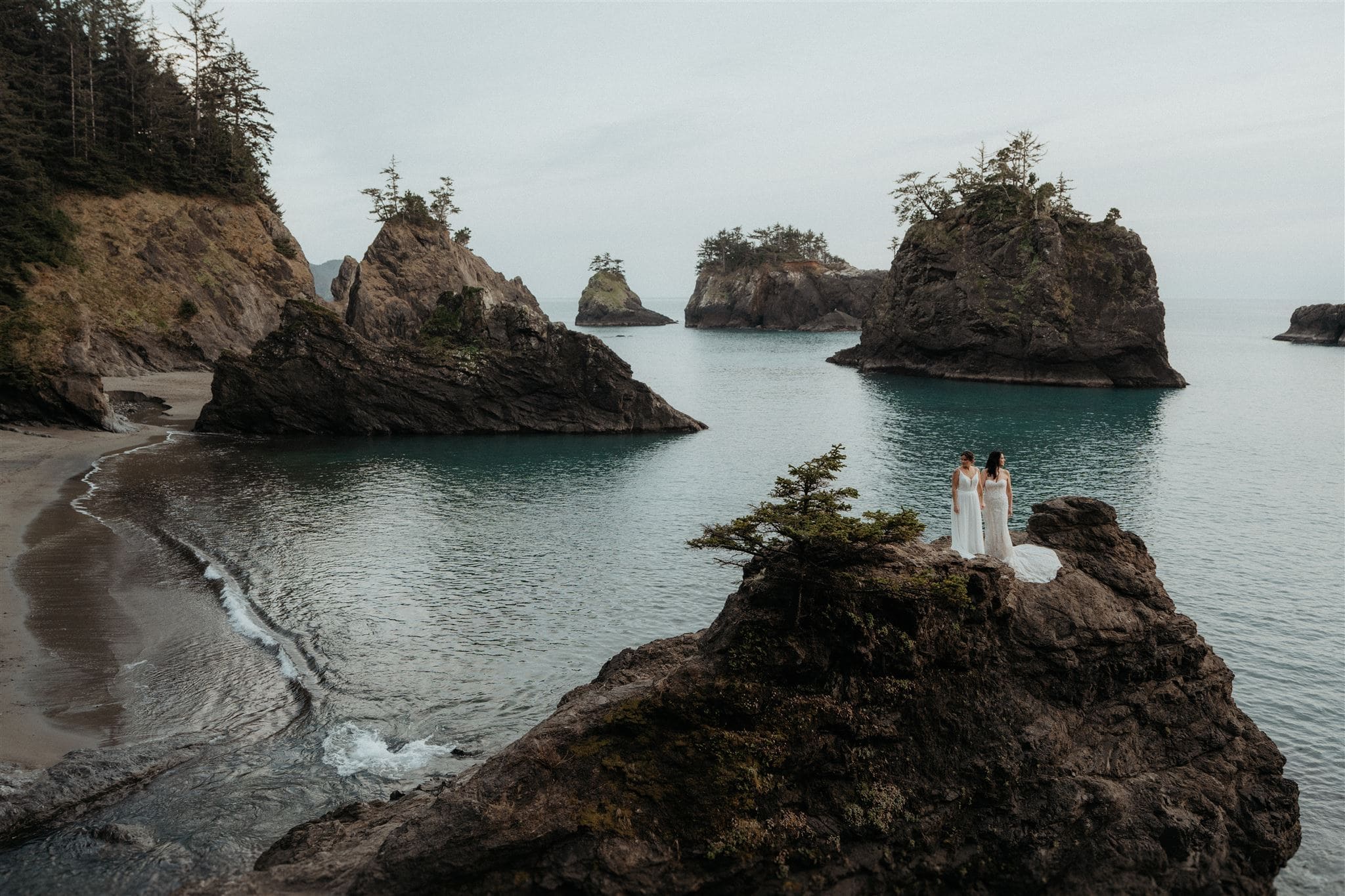 Two brides stand on a rock overlooking the ocean during their Oregon Coast elopement.