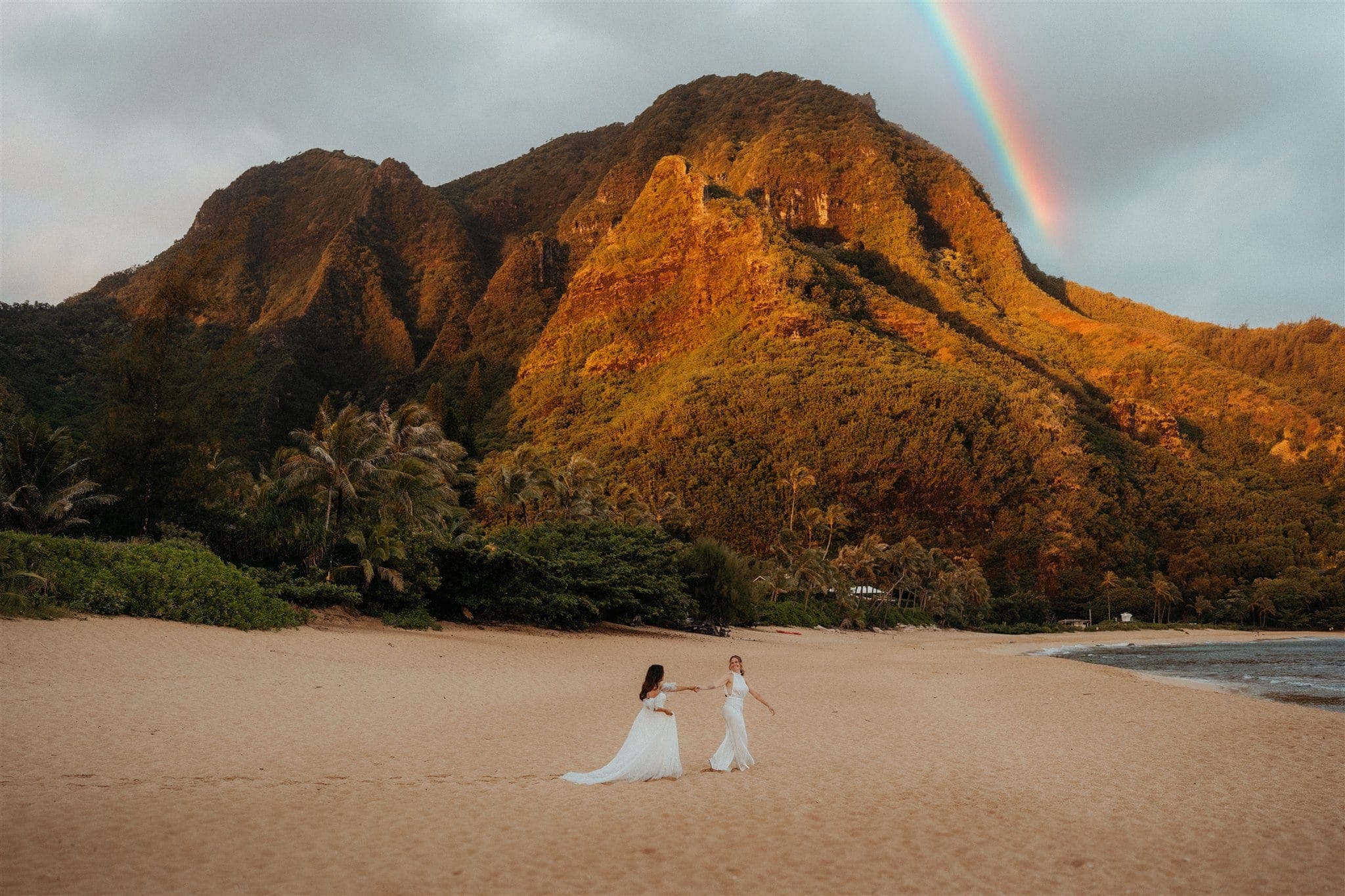 Brides hold hands while walking across Tunnels Beach at sunrise during their Kauai micro wedding.