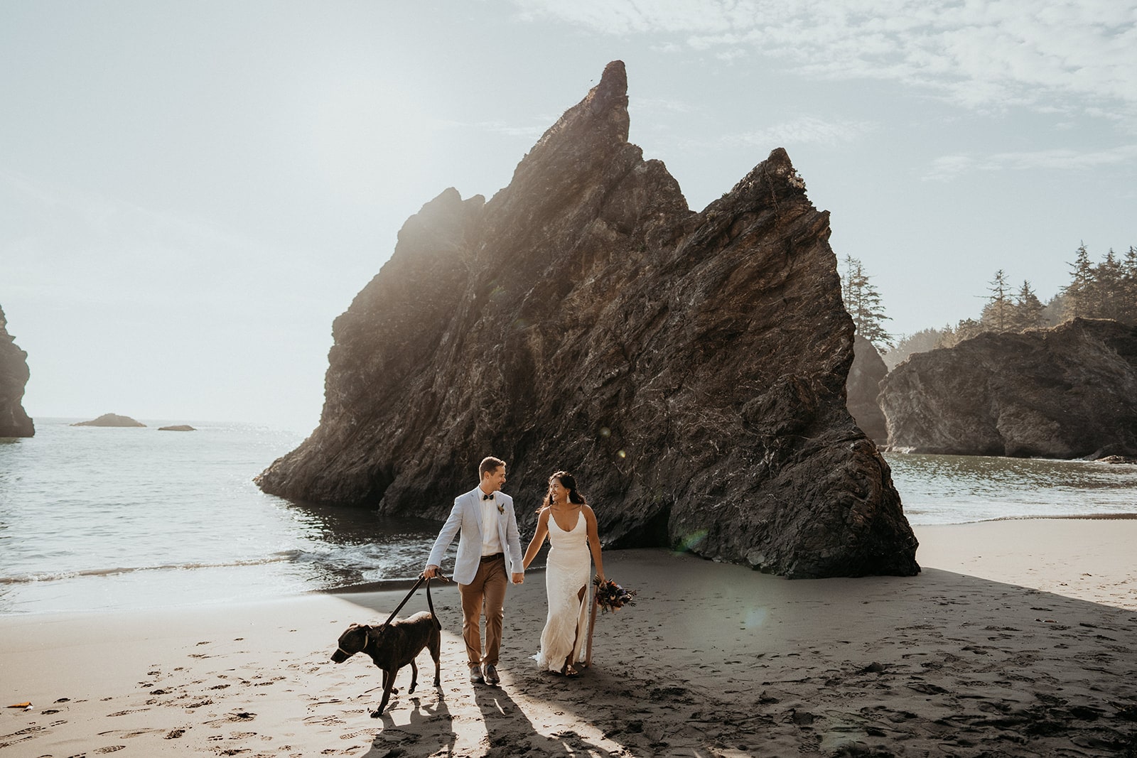 Bride and groom hold hands as they walk their dog on the beach during their Oregon Coast elopement.