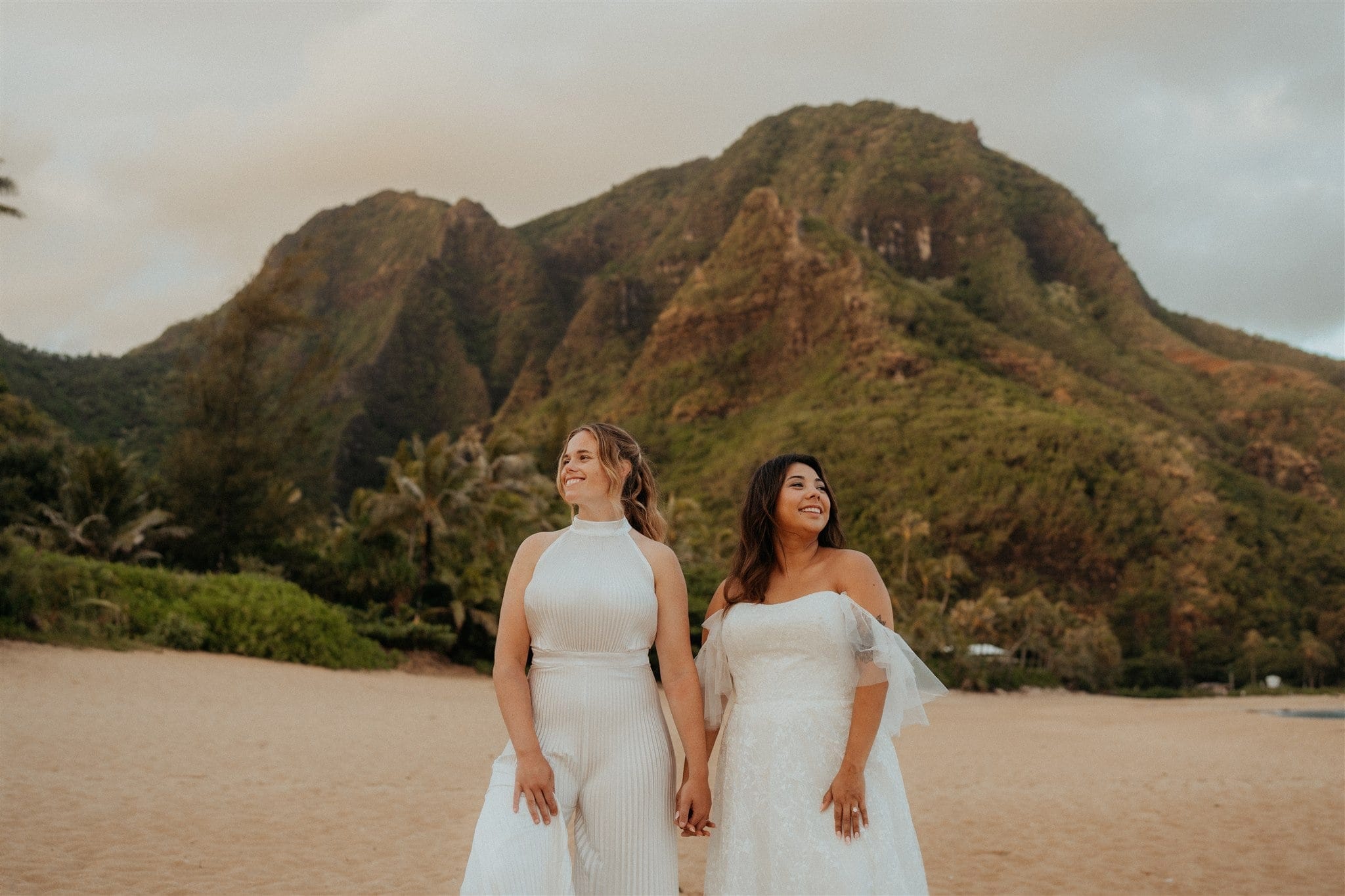 Brides hold hands on Tunnels Beach during their adventure session in Kauai.