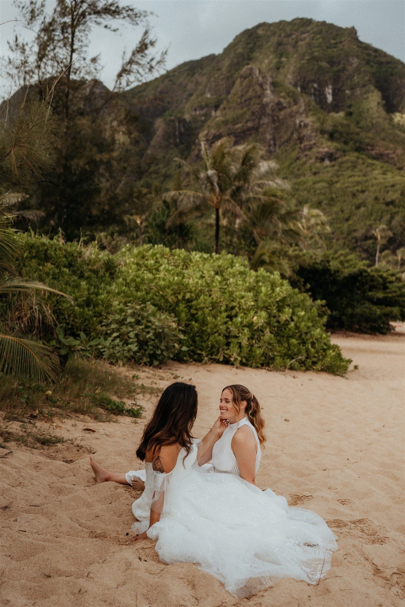 Brides sit together in the sand during their Tunnels Beach adventure photos in Kauai.
