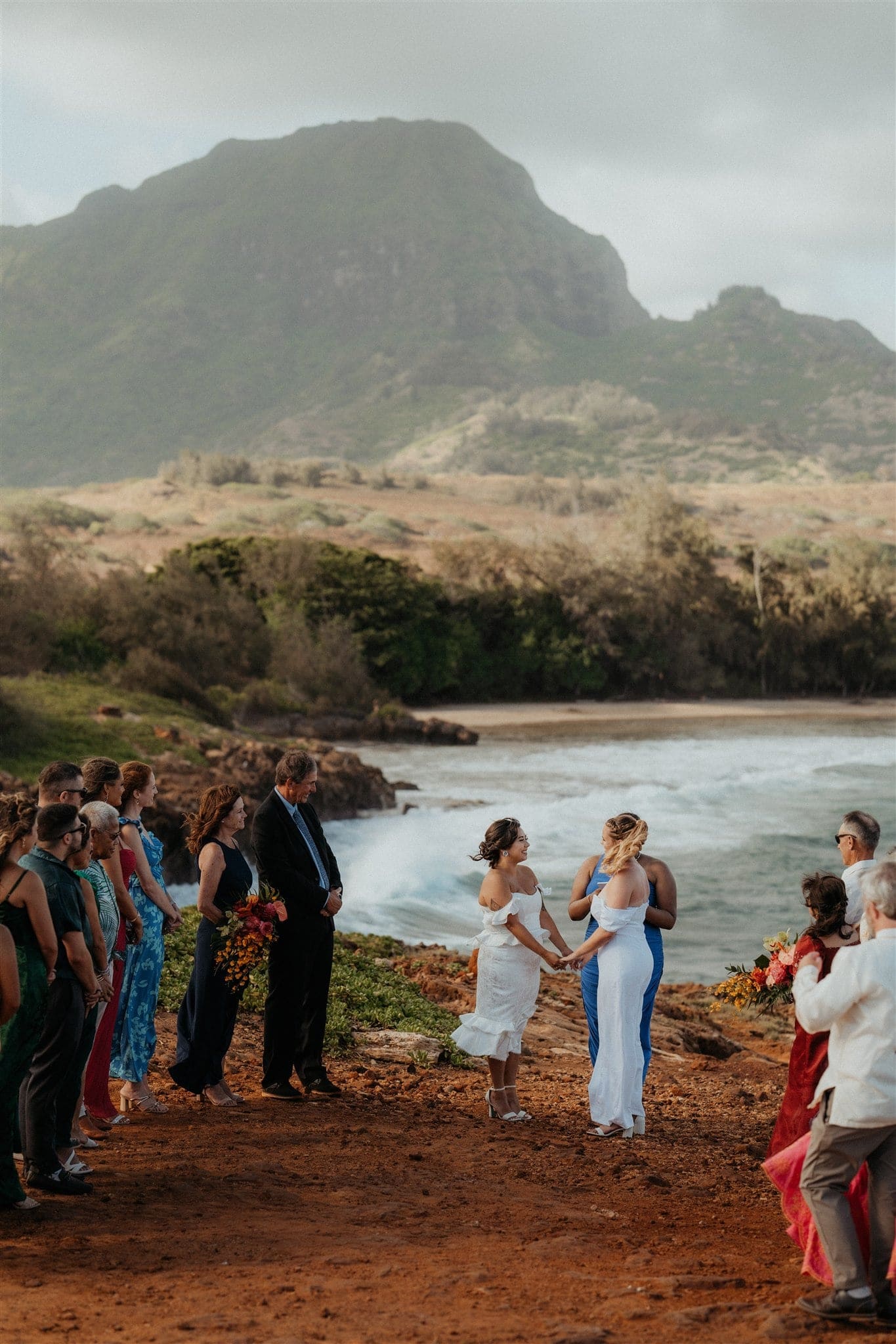 Guests gather around while brides hold hands during their micro wedding ceremony in Kauai on the cliffs.