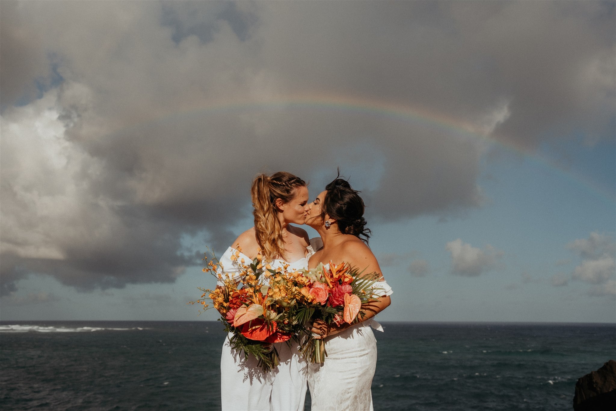 Two brides kiss under a rainbow after their Kauai micro wedding ceremony by the ocean.