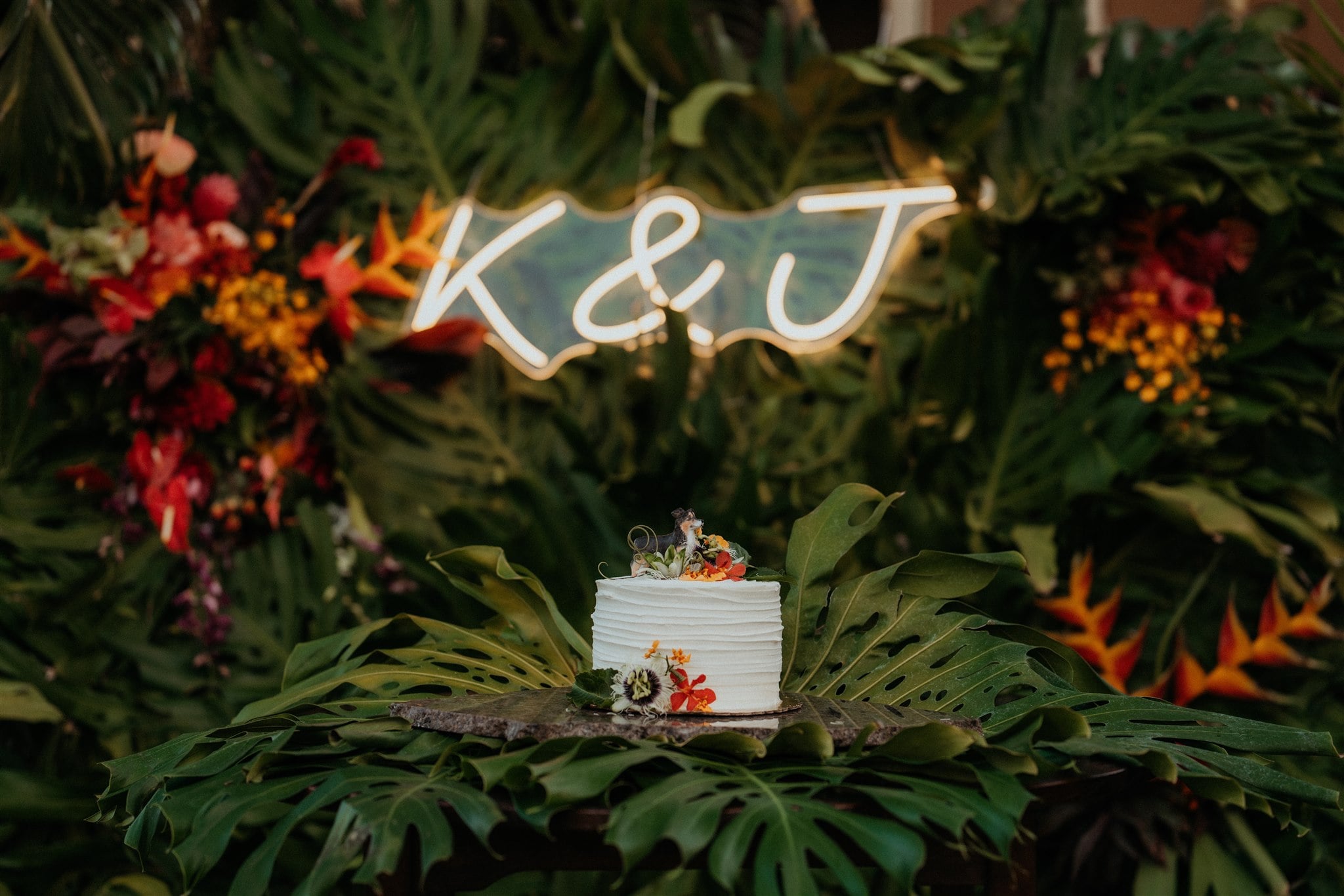 Neon K + J light sits among palm trees with a white frosted wedding cake.