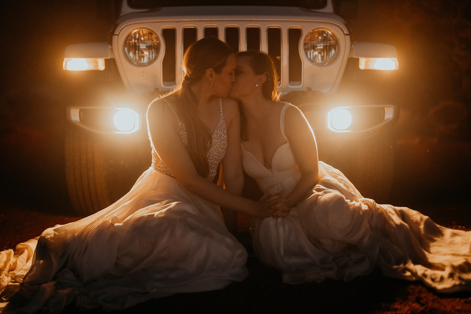 Two brides sit on the ground and kiss in front of a Jeep after dark.