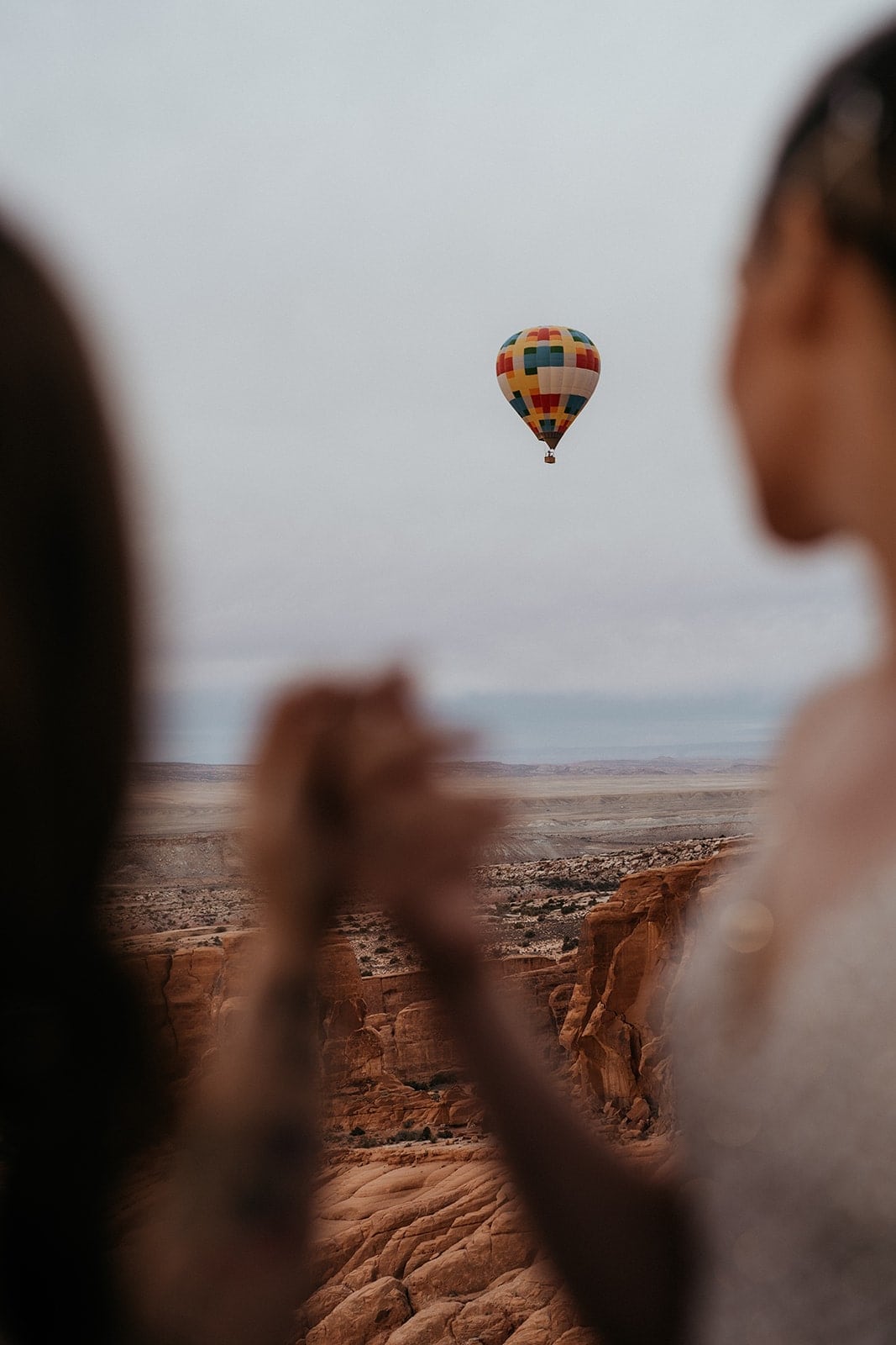 Brides hold hands in a hot air balloon basket and look out at another hot air balloon floating in the sky.