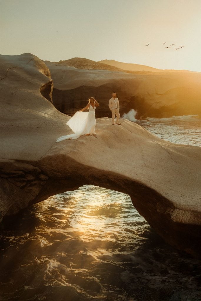 Bride and groom stand on a sandstone beach during their destination wedding in Milos, Greece