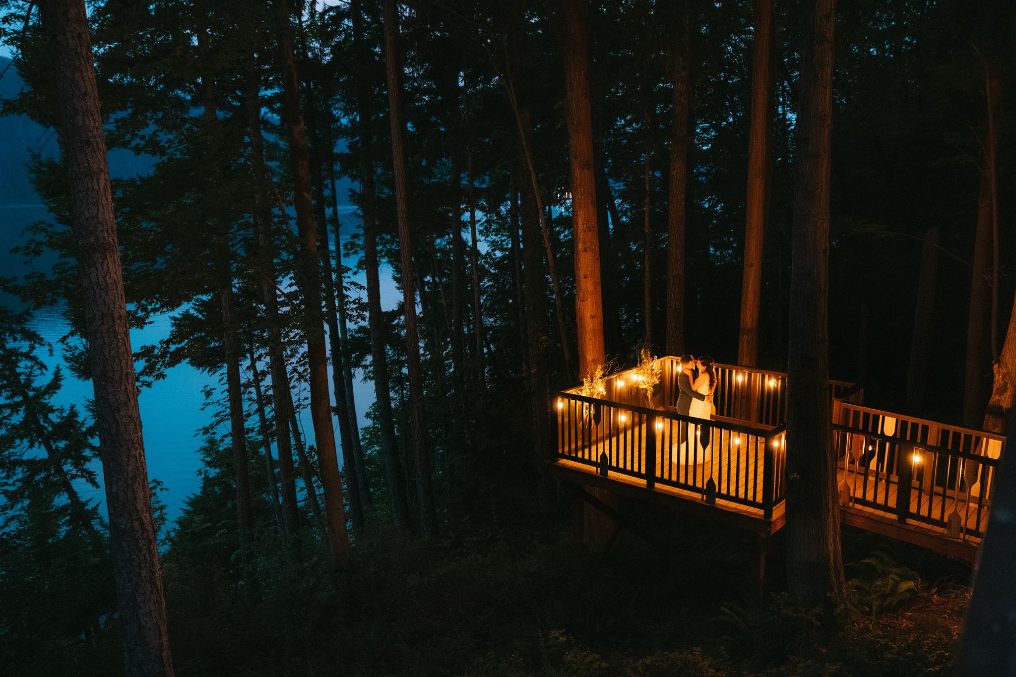 Two brides dance on a balcony decorated with string lights overlooking a lake at the end of their forest elopement day.