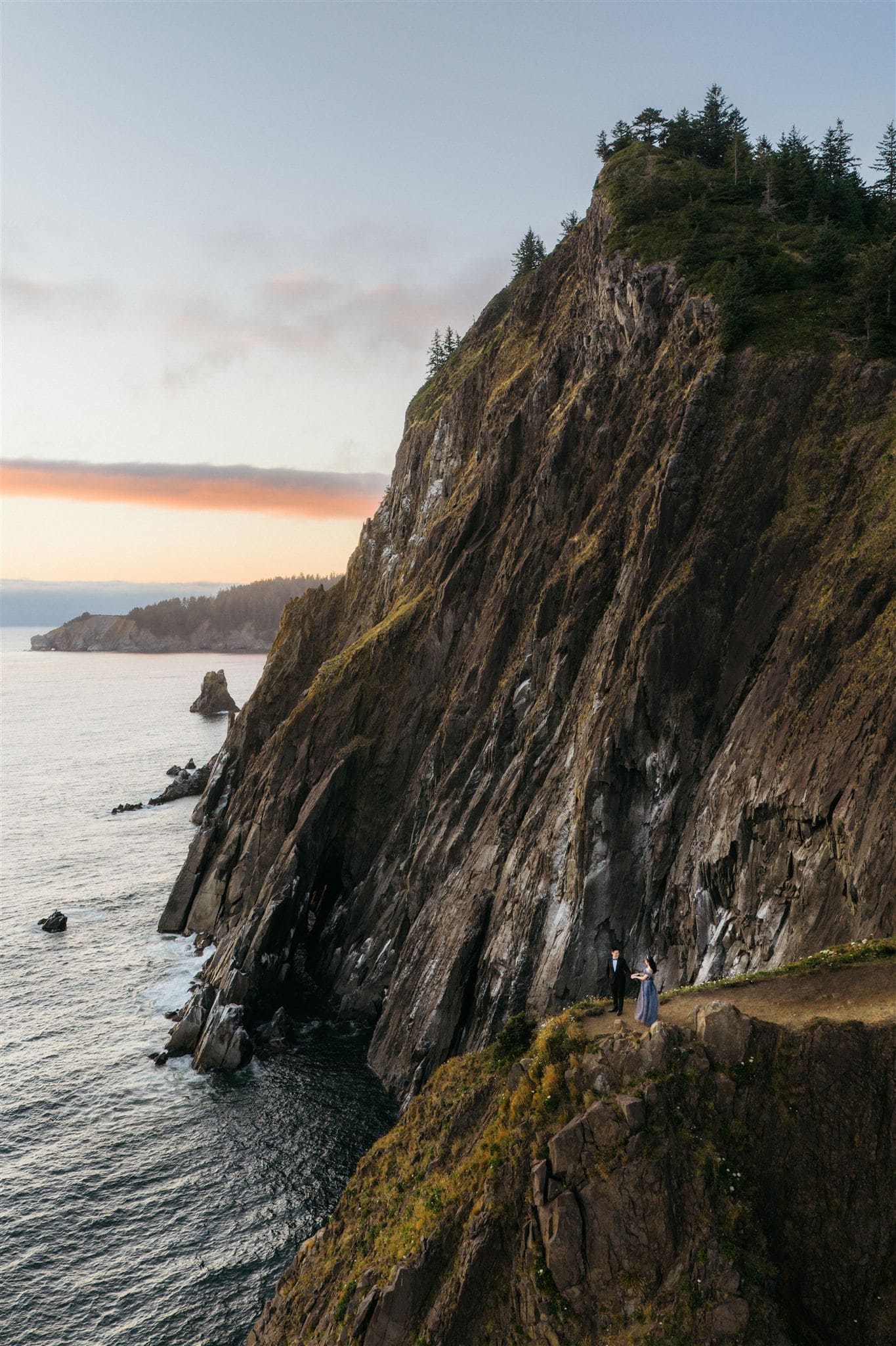 Couple stands on a cliff along the Oregon Coast during sunset.