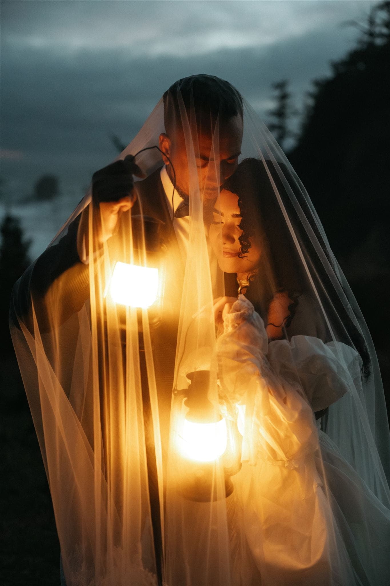 Bride and groom hold lanterns under a wedding veil during their blue hour Oregon Coast portraits.