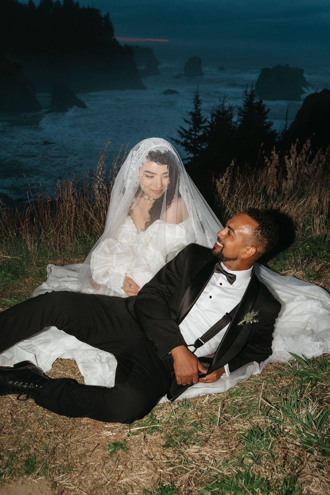 Bride and groom sit on the ground near the Oregon Coast for editorial portraits during blue hour.