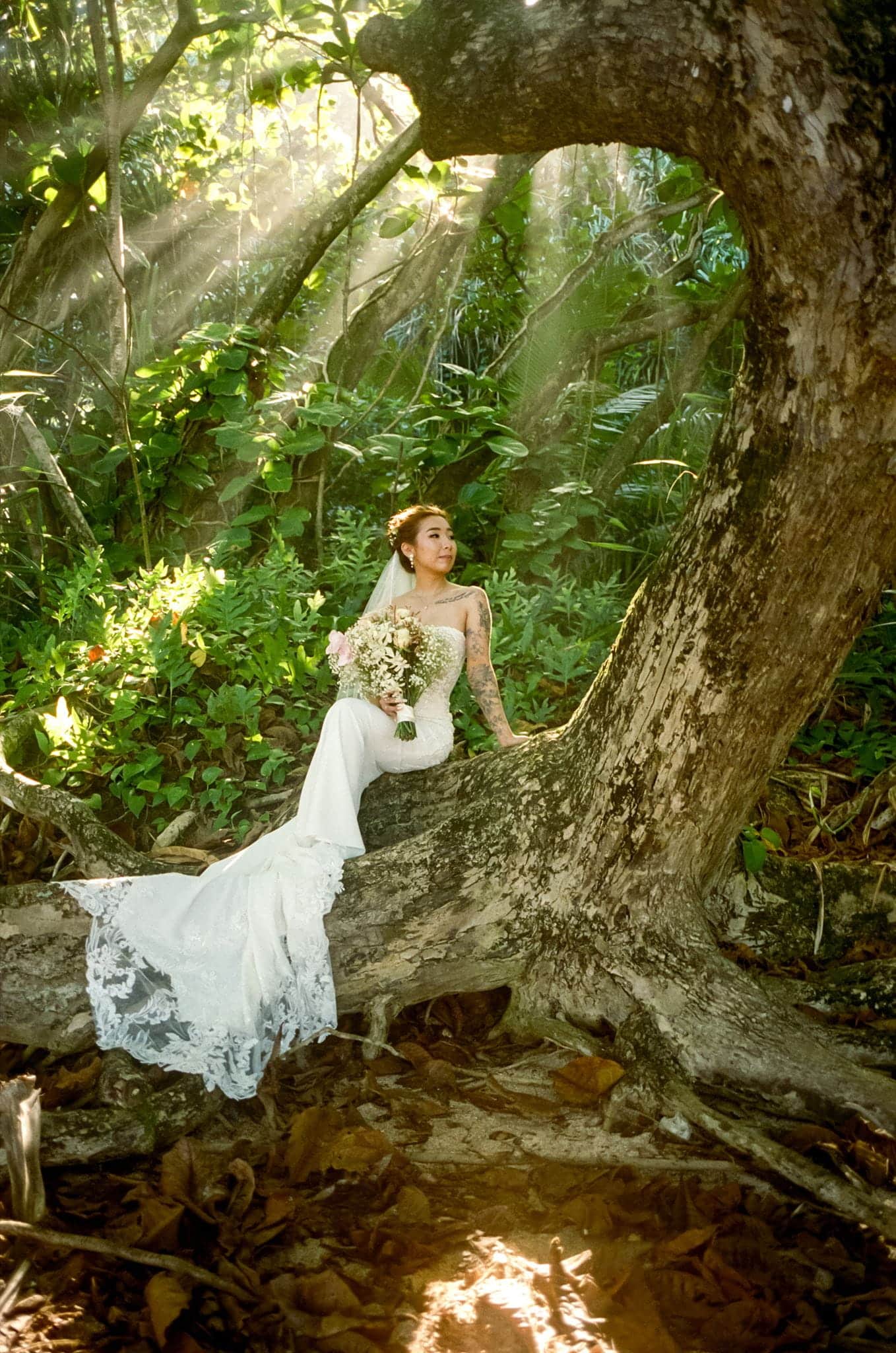 Bride leans against a tree while sunlight filters above during her film bridal portrait session in Hawaii.