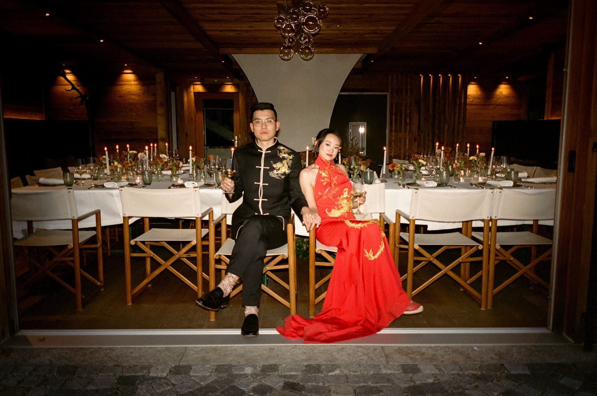 Bride and groom in red and black Asian wedding attire sit at their reception dinner table in the Dolomites, Italy.