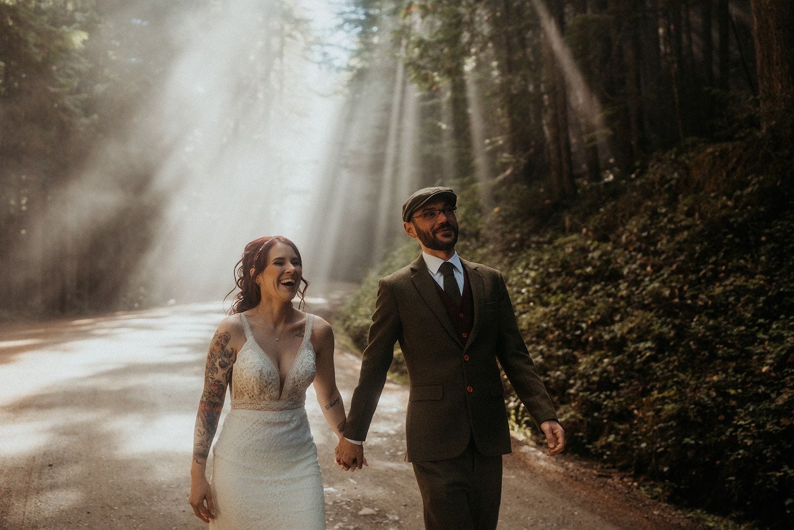 Newlyweds laugh and hold hands while sunlight filters through the forest trees above them.